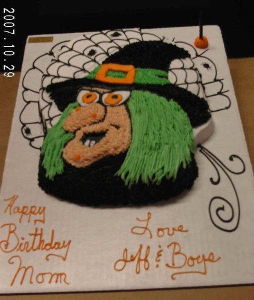 Witch-shaped birthday cake with green hair, black hat, and orange details. Inscription reads 