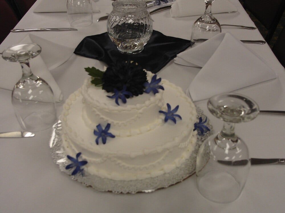 Two-tiered white frosted cake, blue flowers, on a white tablecloth, flanked by upside-down glasses and napkins.