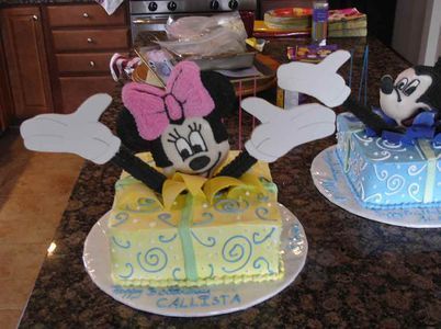 Minnie Mouse cake emerging from a yellow present, with a matching Mickey Mouse cake in the background.