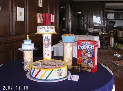 Birthday cakes and cereal box arranged on a table for a celebration, with a dark wood-paneled room in the background.