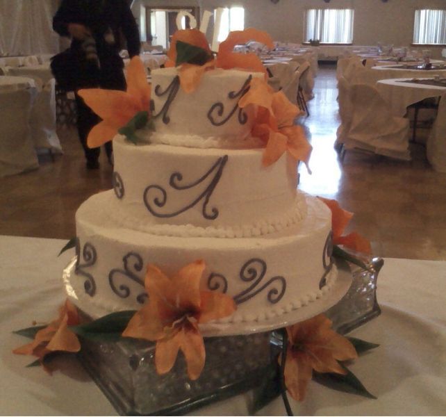 Three-tiered white cake with orange flowers and gray scrollwork on a silver pedestal in a banquet hall.