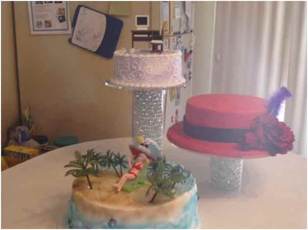 Three decorative cakes on clear stands; a beach scene, a hat, and a lavender-frosted cake.