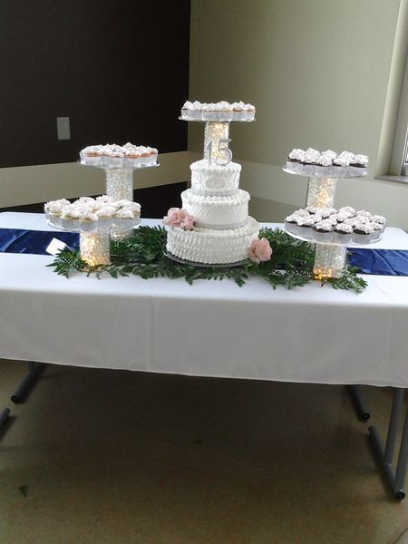 Wedding cake and cupcakes on a white tablecloth, arranged on tiered platforms, with greenery and navy accents.