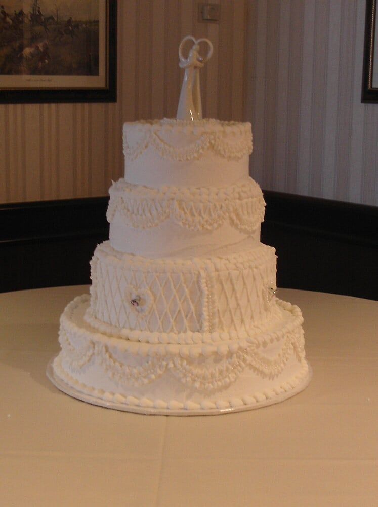 Three-tiered white wedding cake with intricate frosting details and a couple figurine on top, set on a white tablecloth.