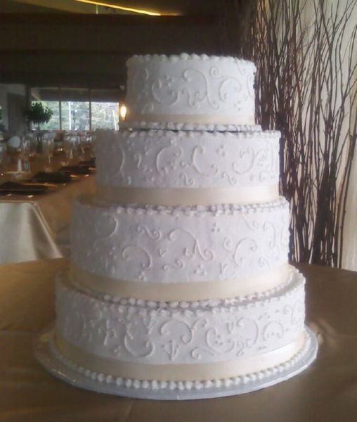 Four-tiered white wedding cake with intricate scrollwork, light gold ribbon, and pearl border, set on a gold tablecloth.