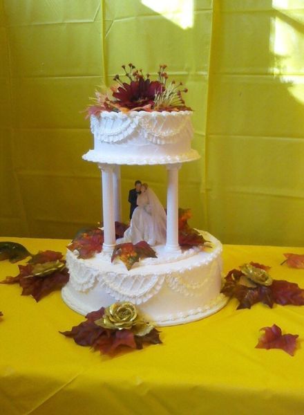 Two-tiered wedding cake with bride and groom figurines, decorated with fall-themed foliage, set on a yellow tablecloth.