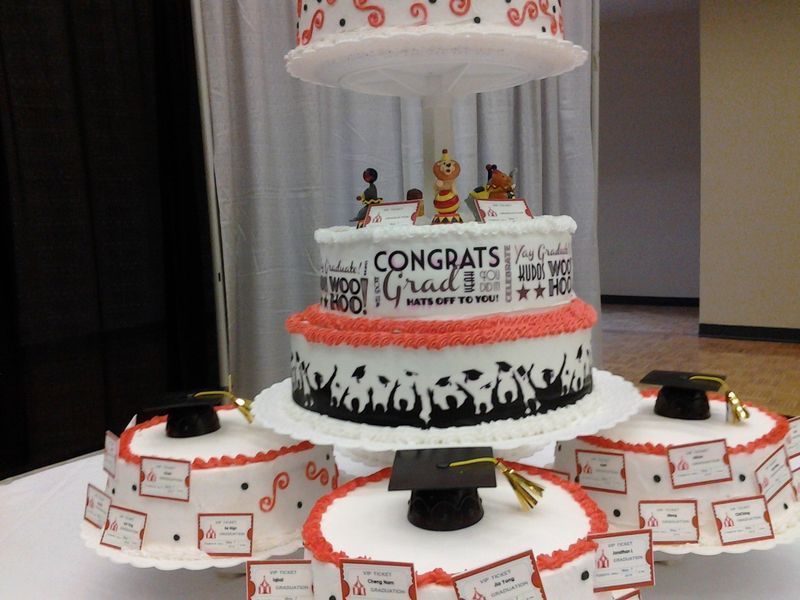 Graduation cake with multiple tiers, topped with figurines and small graduation caps. Red and black accents.
