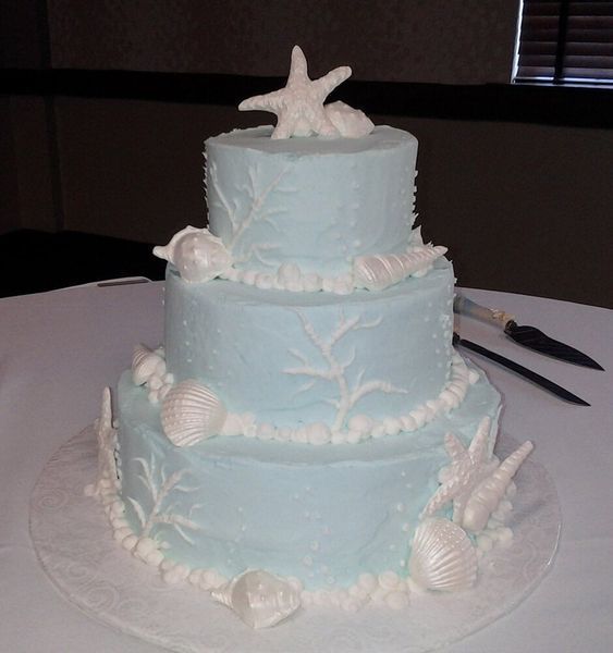Three-tiered blue cake decorated with white seashells, starfish, and coral.