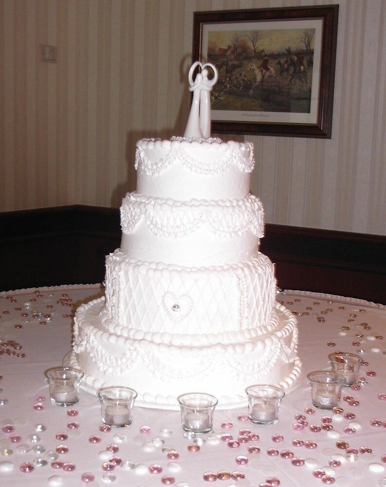 Four-tiered white wedding cake with bride and groom topper, on a table with candles and confetti.