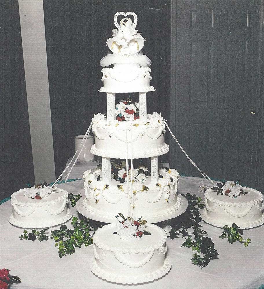 Five-tier white wedding cake with bride and groom topper, surrounded by smaller cakes on a decorated table.