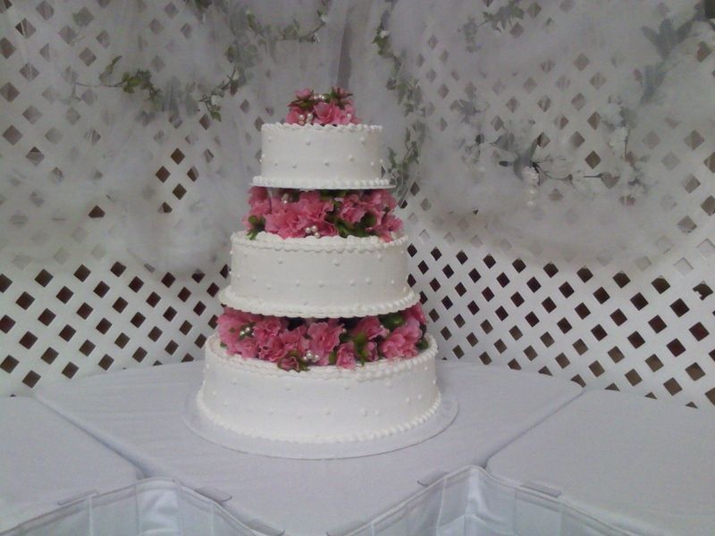 Four-tiered white wedding cake decorated with pink flowers on a white table.