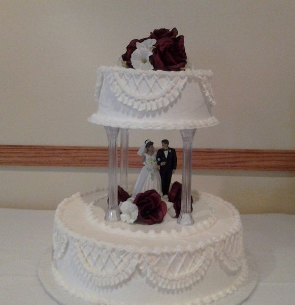 Two-tiered white wedding cake with bride and groom figurines under a gazebo-like top and burgundy floral accents.
