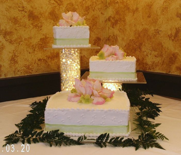 Three-tiered square wedding cake with pink flowers on a white tablecloth, ferns surround the base.