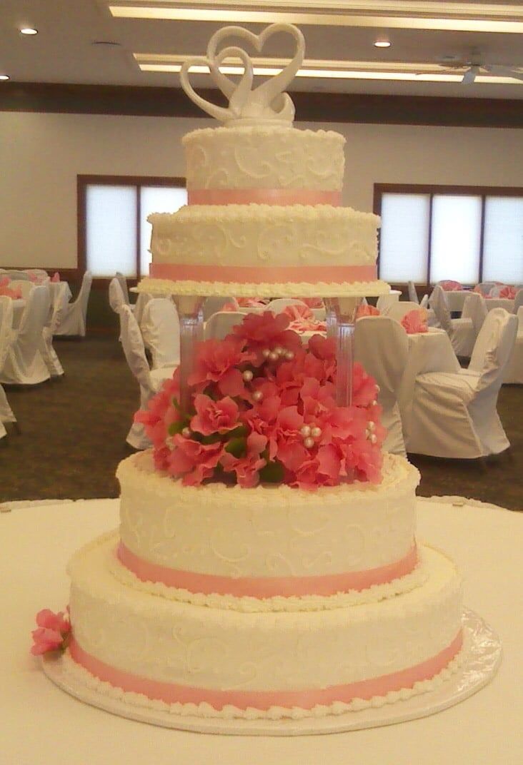 Five-tiered wedding cake with pink ribbon and flowers, topped with swan figures, on a table in a banquet hall.