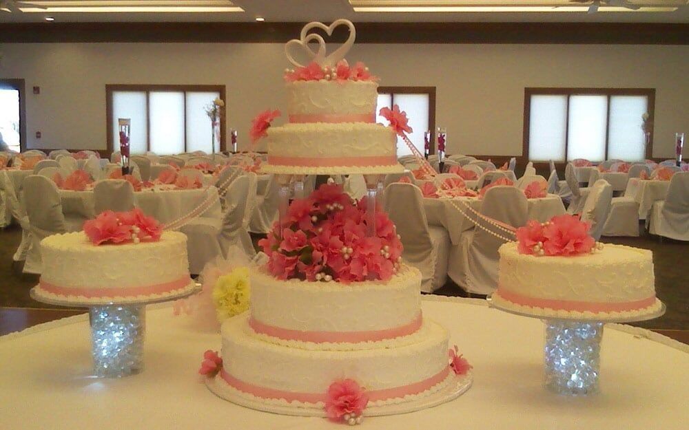 Wedding cake display with tiered cakes, pink flowers, and heart toppers in a banquet hall.