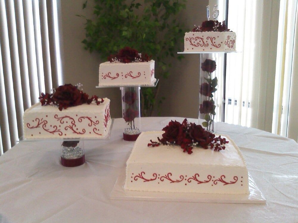 Four-tiered white square cakes with red floral accents and decorative icing on a white table.
