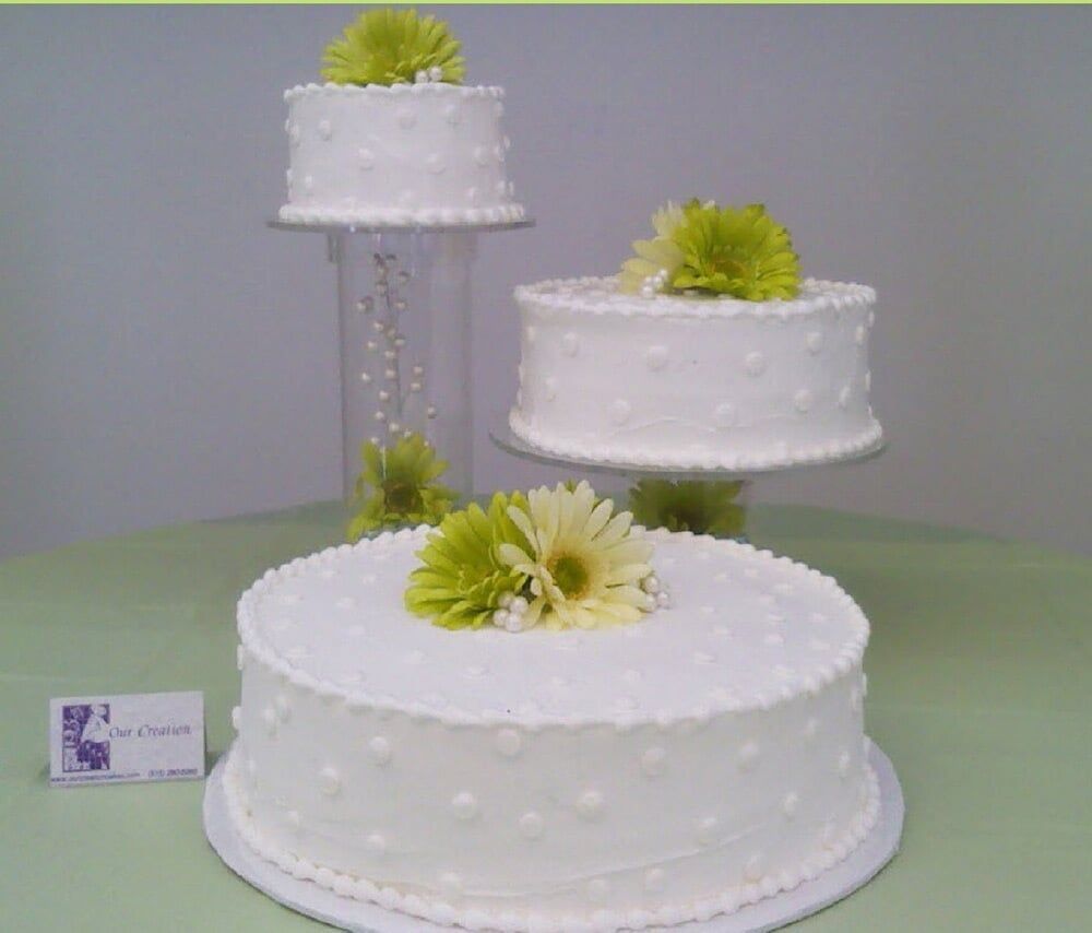 Three-tiered white cake with green flower accents, displayed on stands and a cylinder with pearls, on a green table.