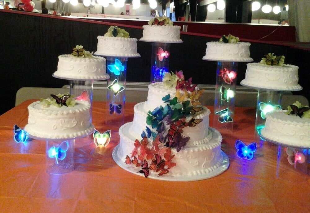 Wedding cakes on clear stands, decorated with butterflies and lit up, set on orange table.