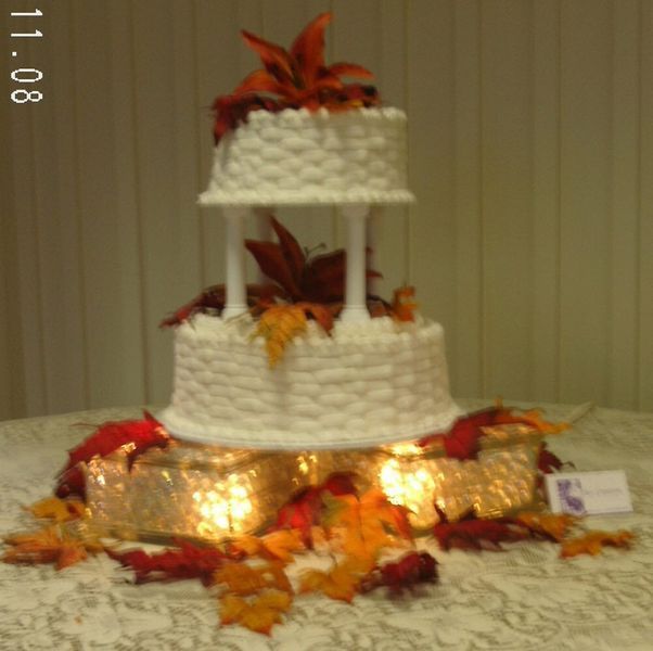Two-tiered white wedding cake with fall leaves and orange flowers, on a lighted base.