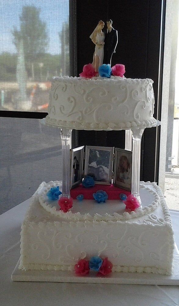 Two-tiered wedding cake with photos and couple figurines, decorated with pink and blue flowers, indoors.