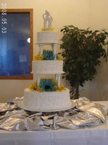 Four-tiered white wedding cake with blue and yellow flowers, topped with a couple figurine.