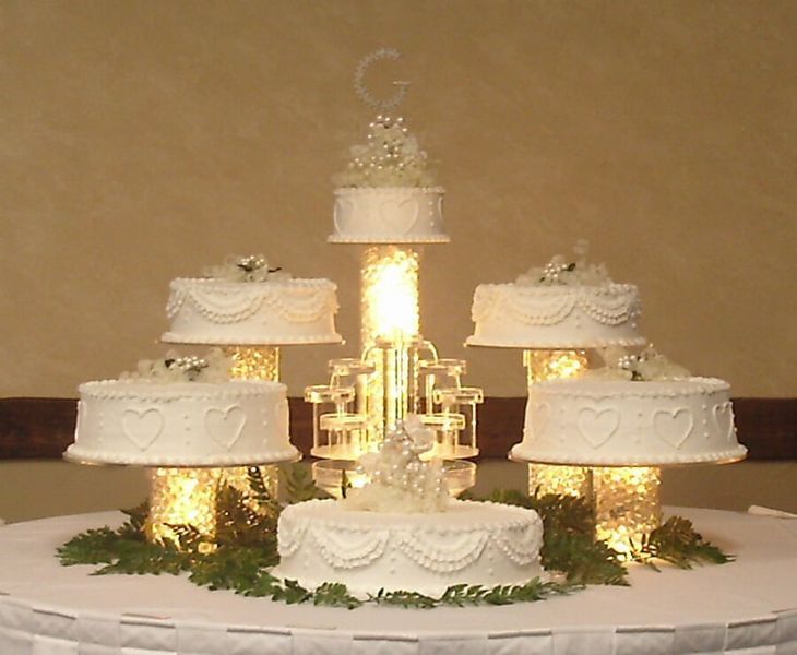 Wedding cake display on a round table, featuring multiple white-frosted cakes on illuminated pedestals, greenery, and a topper.