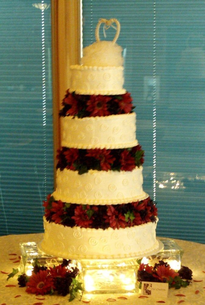 Five-tier wedding cake with red flowers, topped with a teapot. Lit by candles on glass blocks.