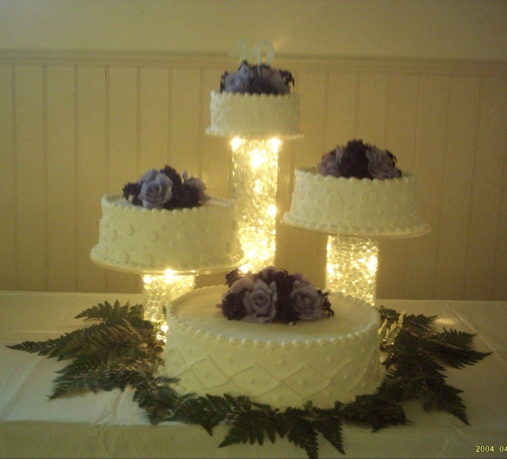 Four-tiered wedding cake with purple floral accents, displayed on illuminated glass pedestals, greenery around the base.