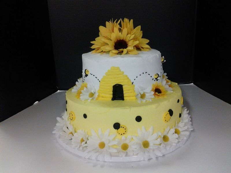 Two-tiered cake decorated with yellow and white frosting, bees, sunflowers, and daisies.