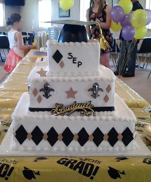 Three-tiered graduation cake with black, white, and gold decorations; balloons and people in the background.
