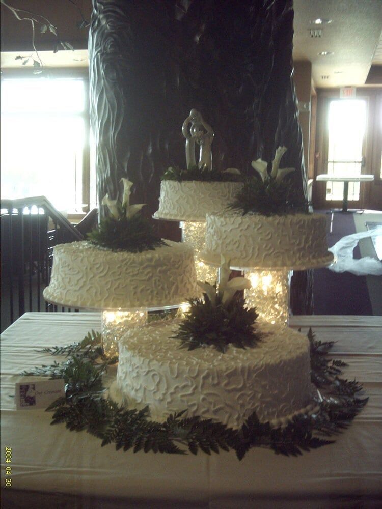 Four-tiered white wedding cake with floral and couple topper, on a table in a room.