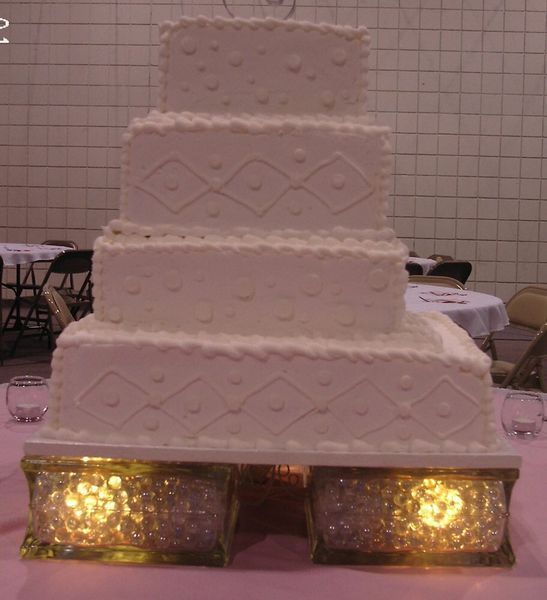Four-tiered white wedding cake decorated with piped details on a lit-up, crystal-filled stand.