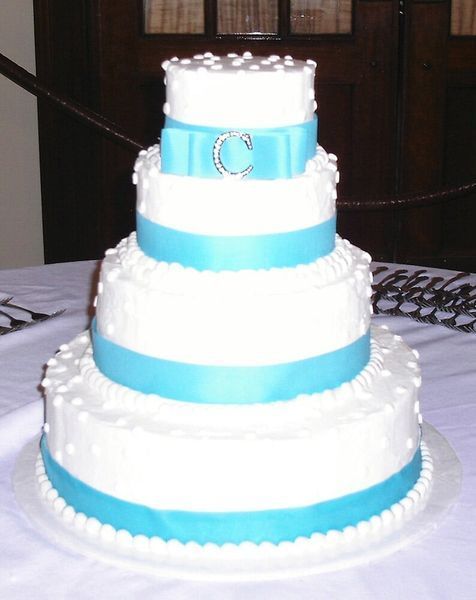 Four-tiered white cake with blue ribbon accents, pearl details, and a jeweled initial 