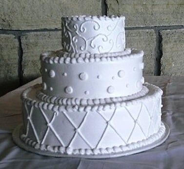 Three-tiered white cake with piped frosting decorations on a white tablecloth.