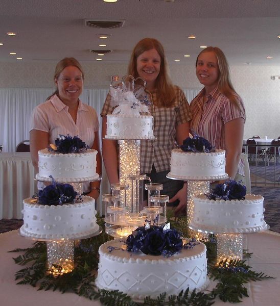 Three women with a tiered wedding cake display, cakes white with blue flowers, indoors.