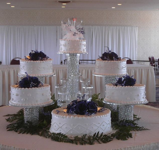 Five-tiered wedding cake display. White cakes with blue flower toppers on crystal pedestals, set on a table.