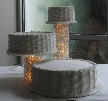 Three cakes with textured, white icing; one on a stand, two on cylinder bases with warm lights.