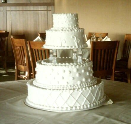 Four-tiered white wedding cake on a table in a dining room, with wood chairs and a white tablecloth.