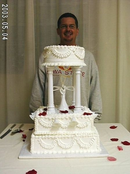 Man smiling behind a tiered wedding cake with decorative columns and red rose petals on a table.