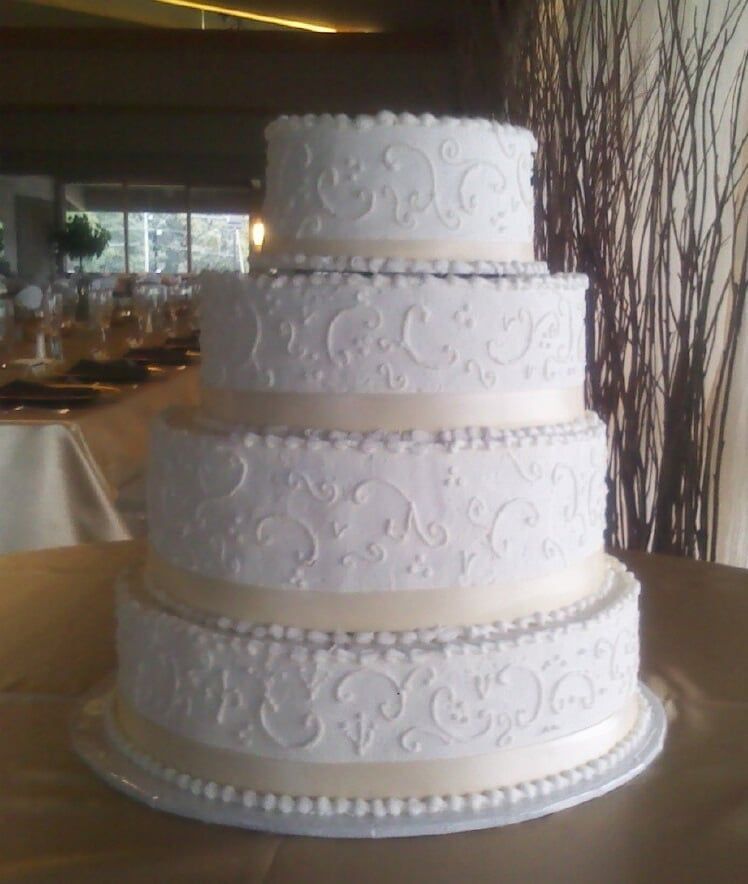 Four-tiered white wedding cake with intricate scrollwork, pearl border, and ribbon accents.