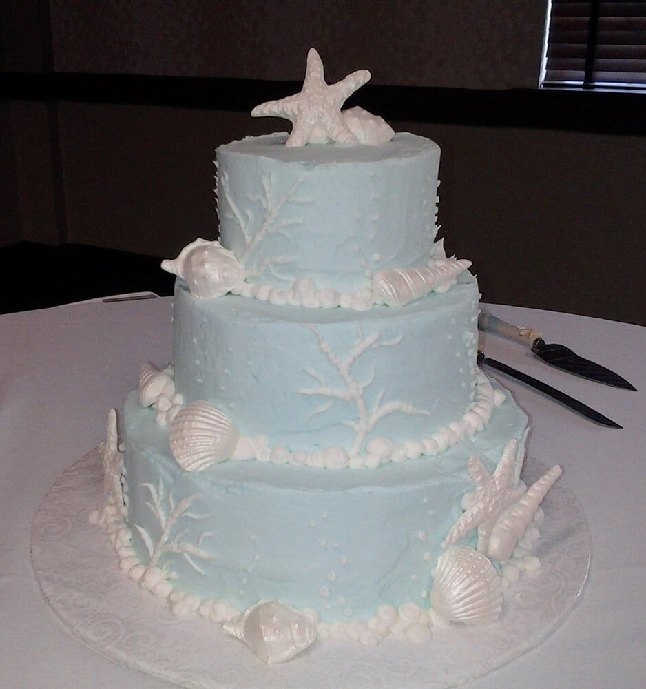 Three-tiered blue cake, decorated with white seashells and starfish, on a white plate.