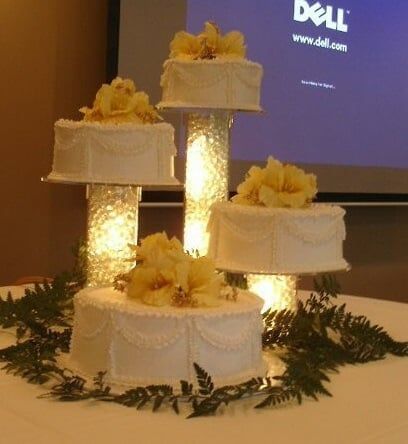 Four-tiered white cake with yellow flower decor, on illuminated glass stands, set on a table with greenery, in front of a projection screen.