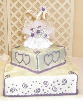 Two-tiered wedding cake, white frosting with purple heart and swirl decorations, topped with a decorative topper.