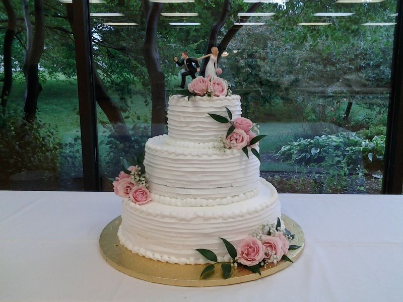 Three-tiered wedding cake with pink roses and figurines; set on a table near a window overlooking greenery.