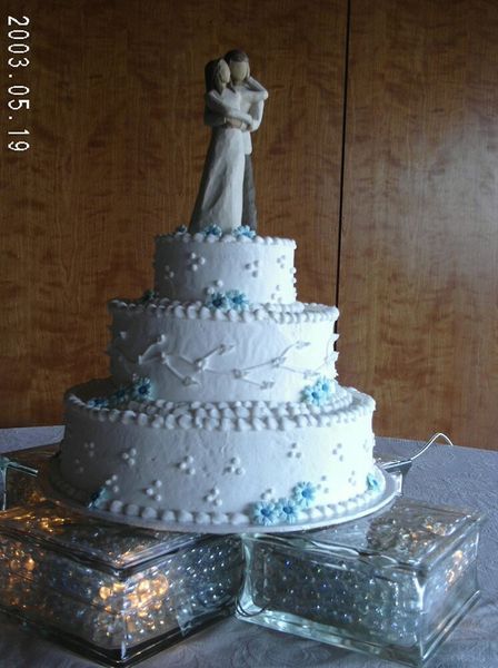 Three-tiered white wedding cake with blue flowers, figurines of a couple embracing on top, on glass blocks.