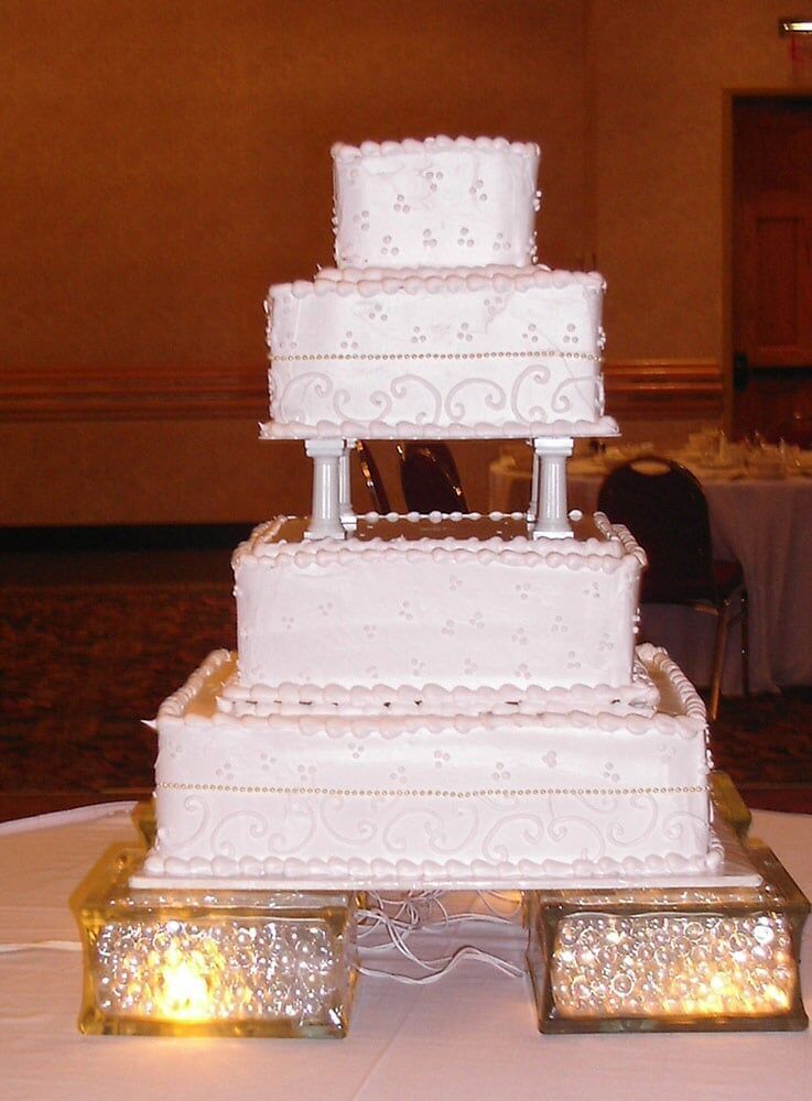 Four-tiered white wedding cake with intricate details, set on a lit, mirrored base in a room.