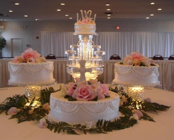 Wedding cake display with four tiers, roses, and greenery on a white tablecloth.