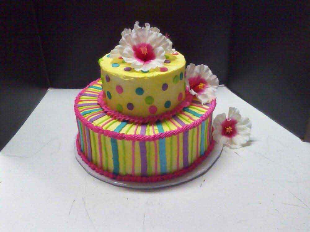 Two-tiered cake with yellow polka dots, striped bottom layer, and hibiscus flower decorations.