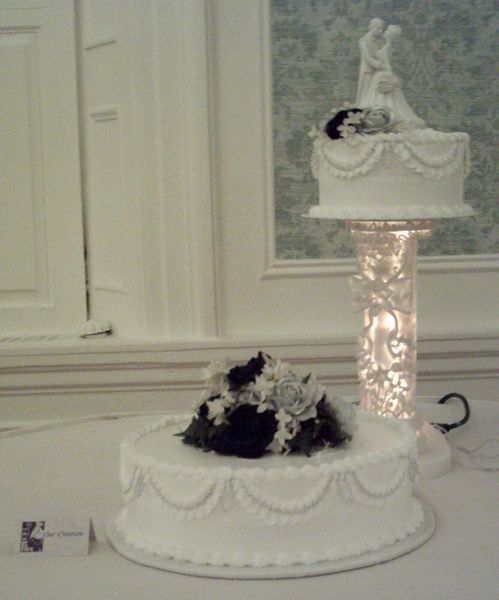 Wedding cake with two tiers, the top tier is raised on a decorated column and topped with bride and groom figurines.