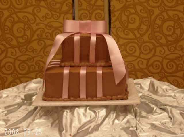 Two-tiered chocolate cake with pink ribbon accents and a bow on top, on a white surface, set against an ornate wall.